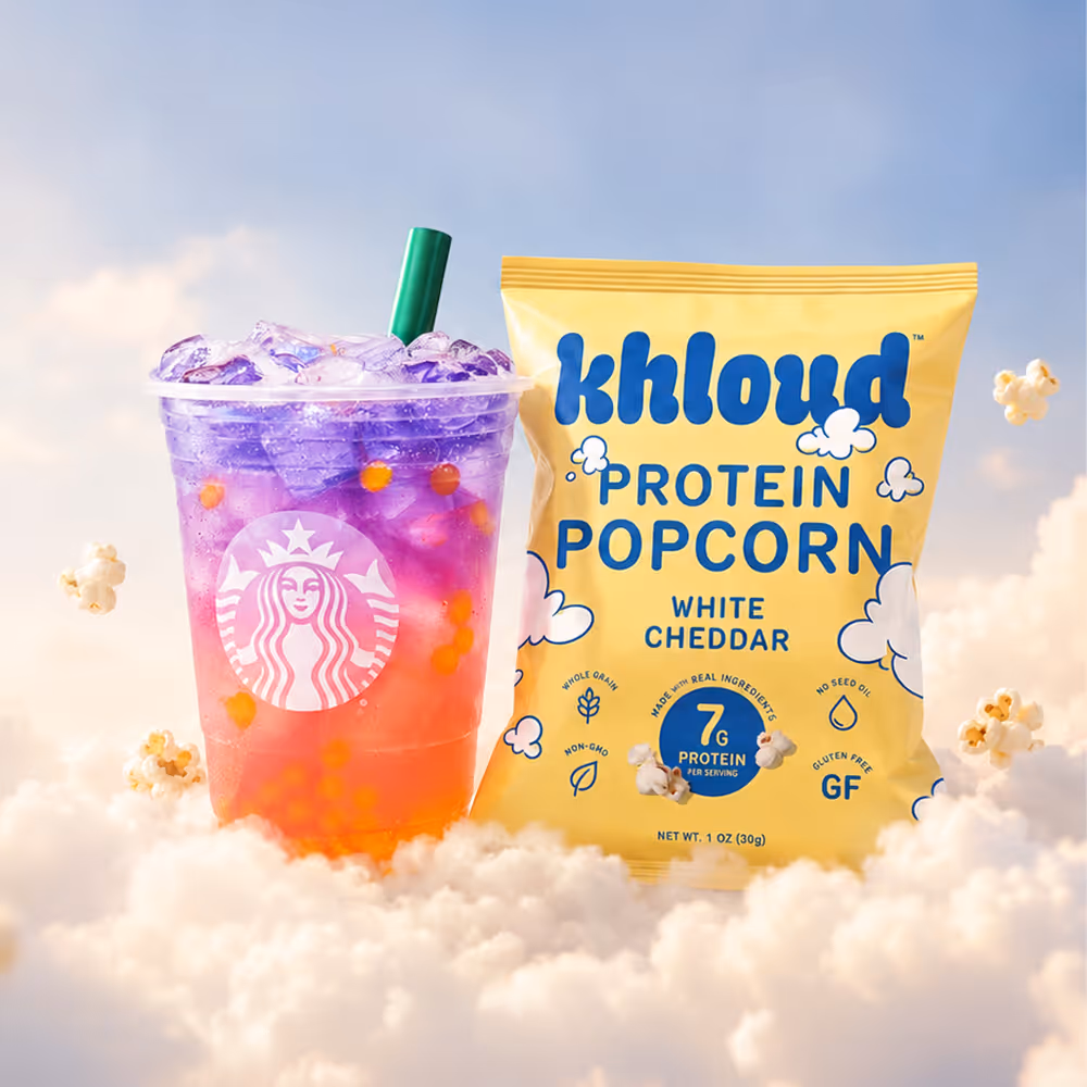 Starbucks Butterfly refresher drink sitting on a cloud in the sky next to Khloud Protein Popcorn White Cheddar in a 1oz bag, surrounded by floating pieces of popcorn